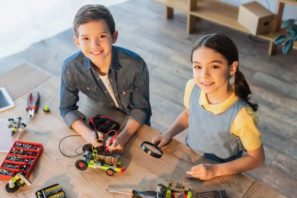 Vista de ángulo alto de los niños con multímetro y lupa ensamblando el modelo de robótica y sonriendo a la cámara - foto de stock