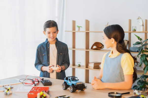 Niño feliz sosteniendo el mando a distancia cerca del modelo de coche hecho a mano y chica en casa - foto de stock