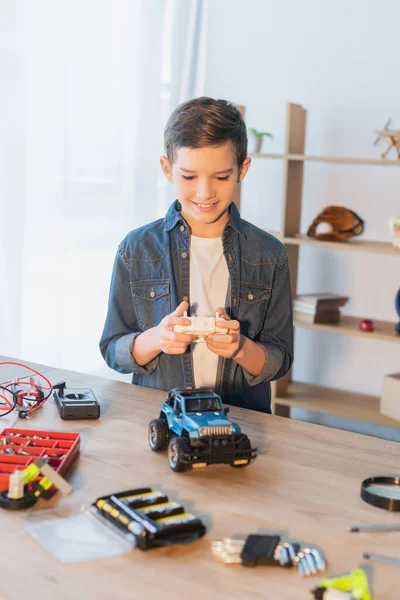 Niño feliz sosteniendo el controlador remoto cerca del modelo de coche hecho a mano en la mesa en casa - foto de stock