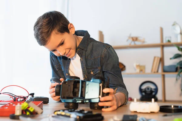 Glücklicher Junge hält handgemachtes Automodell in der Nähe verschwommener Teile auf dem Tisch — Stockfoto