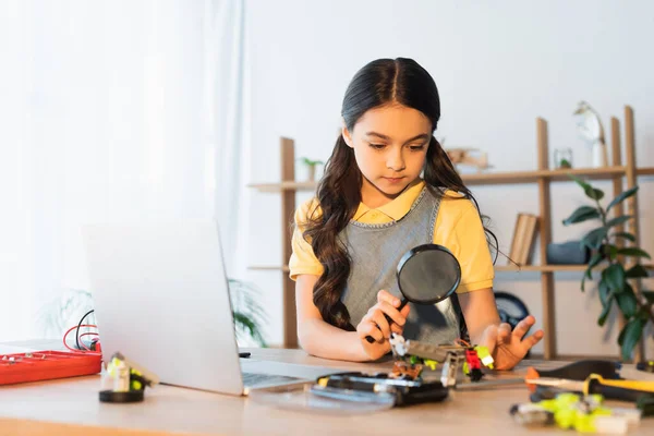 Brunette girl with magnifying glass near laptop and blurred parts of robotics model — Stock Photo