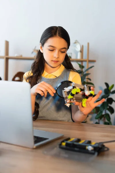 Menina pré-adolescente olhando para o modelo de robótica através de lupa perto de laptop borrado — Fotografia de Stock