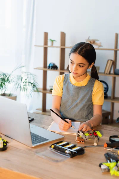 Frühchen schaut auf Laptop und schreibt in Notizbuch neben Details des Robotermodells — Stockfoto