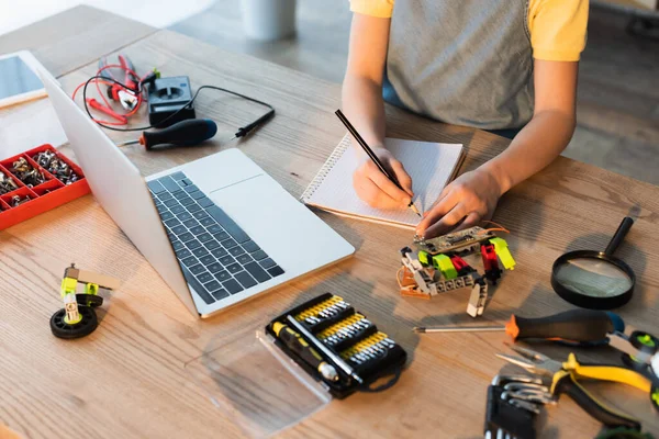 Vista de ángulo alto de la escritura chica recortada en portátil cerca de ordenador portátil y partes mecánicas del modelo de robótica - foto de stock