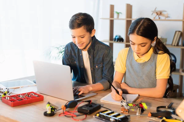 Menino feliz usando laptop enquanto menina escrevendo no notebook perto de detalhes do modelo de robótica — Fotografia de Stock