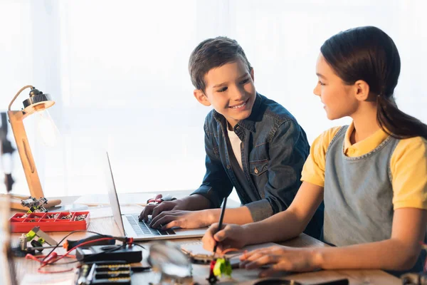 Niño sonriente usando el ordenador portátil mientras mira a la chica escribiendo cerca de partes borrosas del modelo de robótica - foto de stock