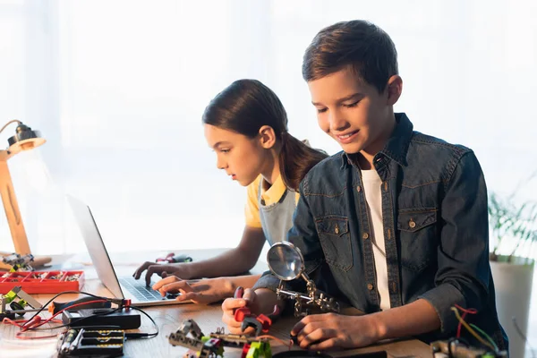 Menino sorridente com lupa montagem robótica modelo perto da menina usando laptop — Fotografia de Stock