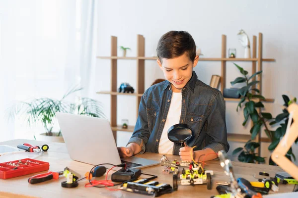 Улыбающийся мальчик держит увеличительное стекло рядом с частями модели робота и ноутбука — стоковое фото