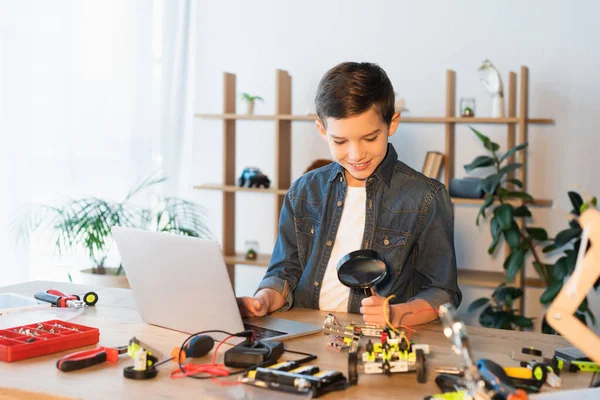 Menino sorrindo segurando lupa perto de partes do modelo de robótica e laptop — Fotografia de Stock