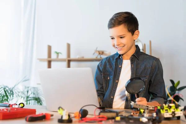 Lächelnder Junge mit Lupe und Blick auf Laptop in der Nähe verschwommener mechanischer Teile auf dem heimischen Tisch — Stockfoto