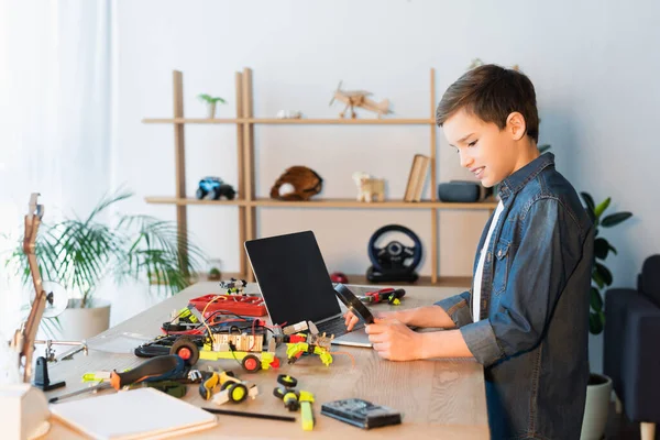 Side view of kid with magnifying glass near details of robotics model and laptop with blank screen — Stock Photo