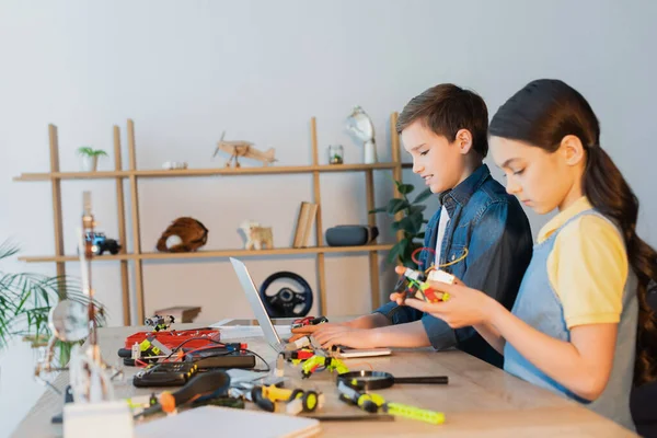 Preadolescente chico usando laptop cerca chica celebración mecánica parte de robotics modelo - foto de stock