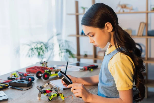 Vista laterale di ragazza con lente di ingrandimento scrittura in notebook vicino a parti meccaniche del modello di robotica — Foto stock