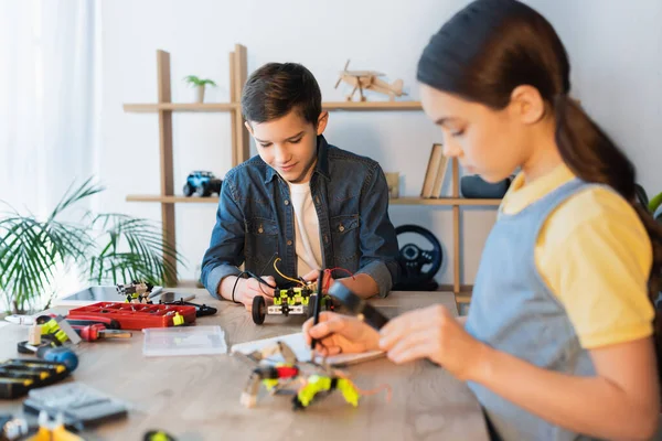Boy assembling robotics model near blurred girl with magnifier writing in notebook — Stock Photo