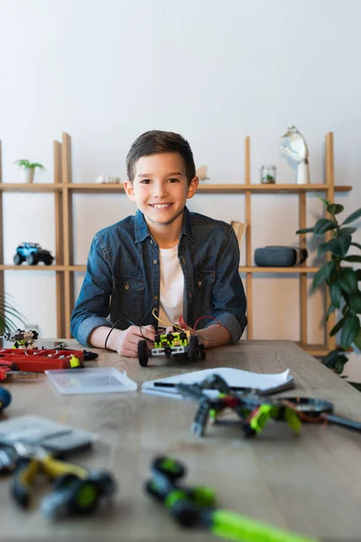 Веселый мальчик смотрит в камеру рядом с механическими деталями робототехники модели на столе у себя дома — стоковое фото
