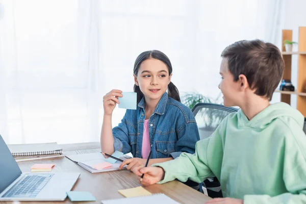 Sonriente chica sosteniendo vacío pegajoso nota cerca borrosa chico y portátil en el escritorio - foto de stock