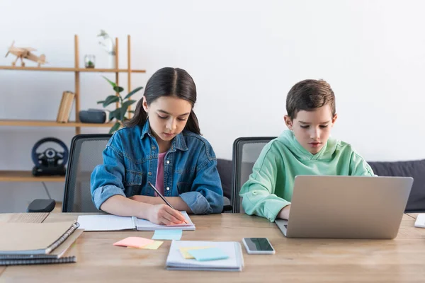 Мальчик с помощью ноутбука и девушка, пишущие в ноутбуке рядом со смартфоном с пустым экраном на столе дома — стоковое фото