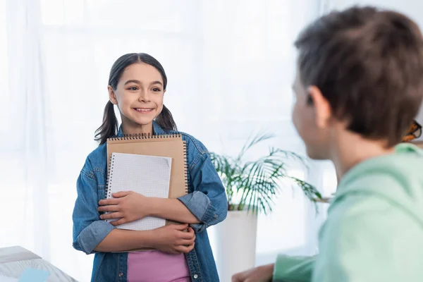 Cheerful girl holding notebooks and looking at blurred boy at home — Stock Photo