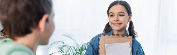 Sonriente chica con copybooks mirando borrosa chico en casa, pancarta - foto de stock