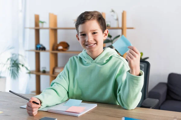 Niño feliz sosteniendo nota adhesiva vacía y lápiz cerca de copybook en el escritorio en casa - foto de stock