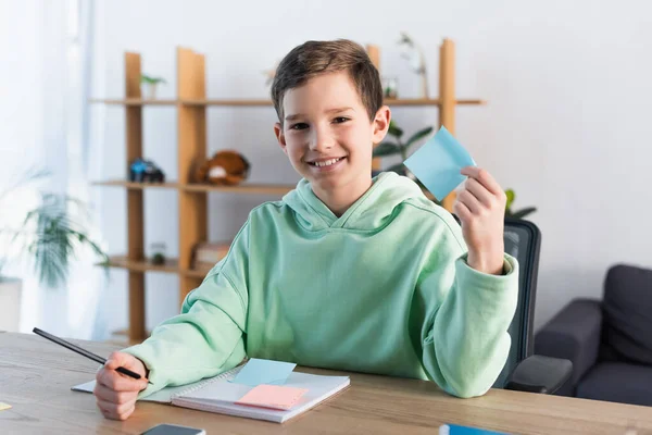 Menino feliz segurando nota pegajosa vazia e lápis perto copybook na mesa em casa — Fotografia de Stock