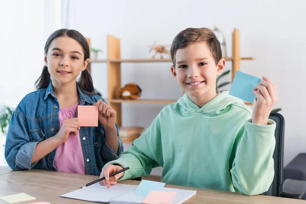 Enfants souriants regardant la caméra et montrant des notes collantes vierges — Photo de stock