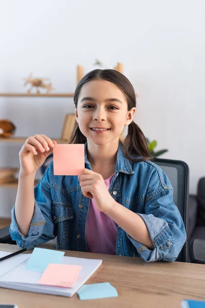 Cheerful girl showing empty sticky note and looking at camera at home — Stock Photo