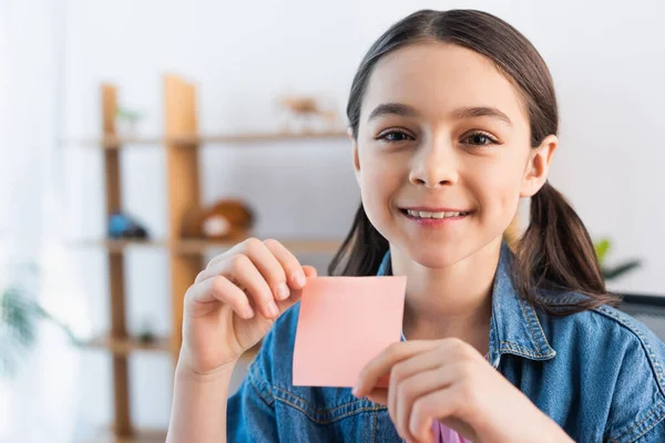 Happy brunette girl looking at camera while holding blank sticky note — Stock Photo