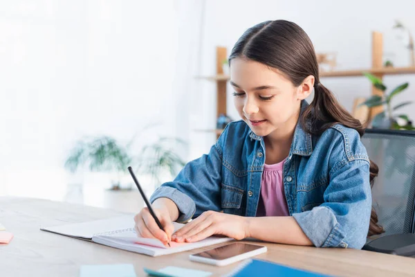 Chica morena sonriente escribiendo en un cuaderno mientras hace la tarea cerca del teléfono celular con pantalla en blanco - foto de stock