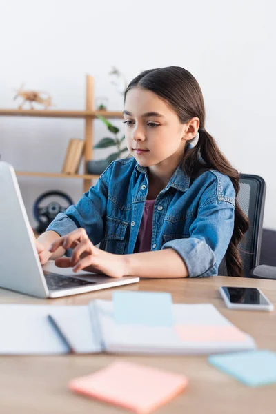 Focused schoolgirl typing on laptop while learning at home near smartphone with blank screen — Stock Photo