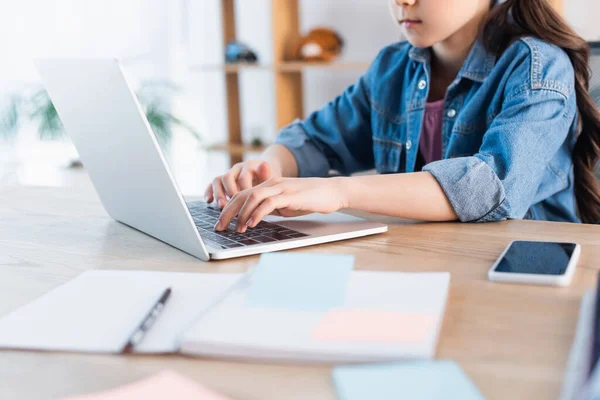 Обрізаний вид дівчини, що друкує на ноутбуці біля розмитої копії під час навчання вдома — стокове фото