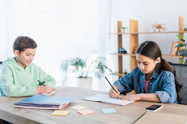 Lächelnder Junge und konzentriertes Mädchen, die in Notizbücher neben leeren Haftnotizen schreiben — Stockfoto