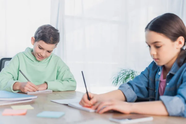 Preteen kids writing in notebooks while doing homework on blurred foreground — Stock Photo