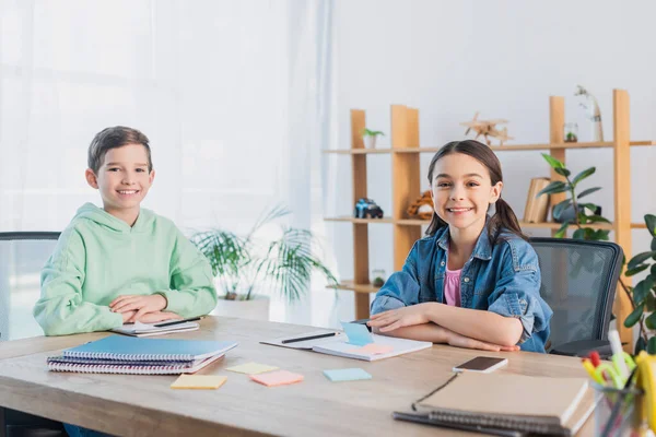 Cheerful kids sitting at desk near notebooks and sticky notes while smiling at camera — Stock Photo