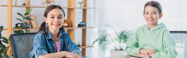 Happy schoolkids looking at camera while sitting at desk at home, banner — Stock Photo