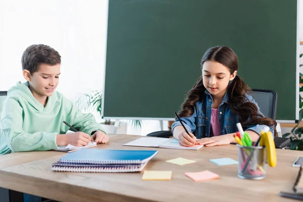 Ragazzo sorridente che scrive nel taccuino vicino alla ragazza e alla lavagna a scuola — Foto stock