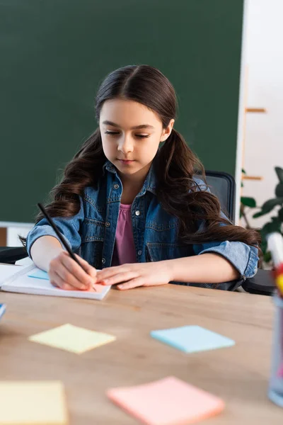 Ragazza bruna che scrive in notebook vicino a note appiccicose sfocate e lavagna a scuola — Foto stock