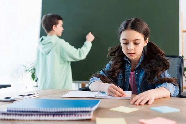 Brünettes Schulmädchen schreibt in Notizbuch in der Nähe Junge an Tafel auf dem Hintergrund — Stockfoto