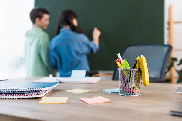 Selective focus of school desk with notebooks and sticky notes near blurred classmates writing on chalkboard — Stock Photo