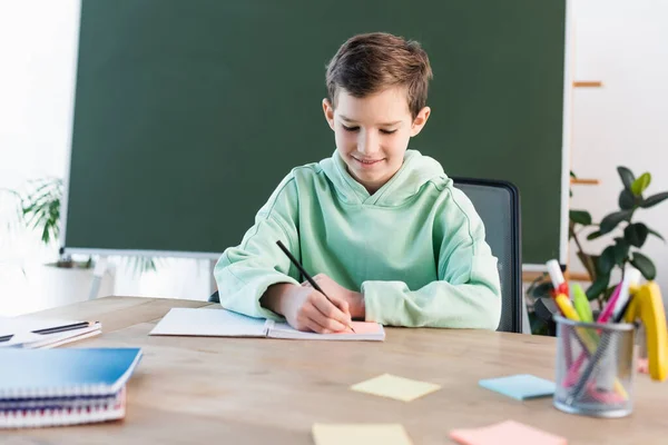 Menino sorridente escrevendo em caderno perto de quadro-negro e notas pegajosas borradas na mesa da escola — Fotografia de Stock