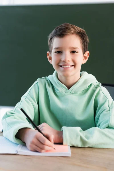 Happy schoolboy writing in notebook and looking at camera in school — Stock Photo