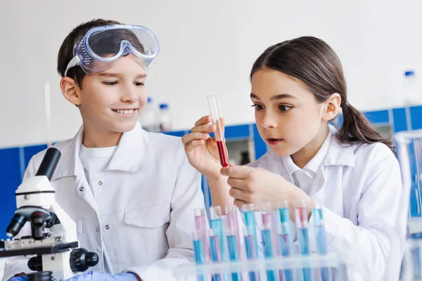 Изумленная девушка держит пробирку с красной жидкостью рядом улыбающийся мальчик и микроскоп в лаборатории — стоковое фото