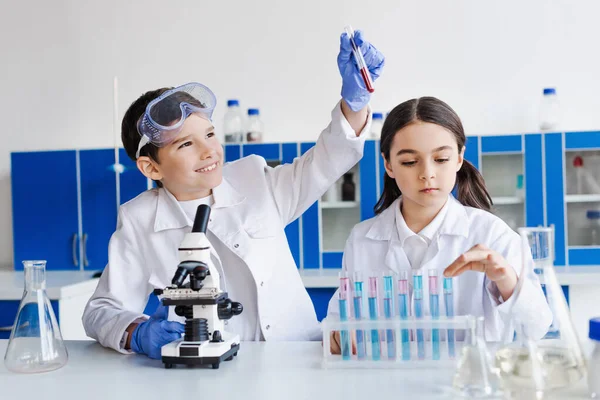 Niño alegre sosteniendo tubo de ensayo con líquido rojo cerca del microscopio y chica seria en laboratorio — Stock Photo