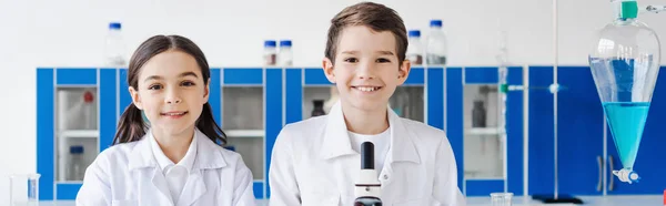 Amigos preadolescentes felices mirando la cámara cerca del frasco con líquido en el laboratorio, pancarta - foto de stock