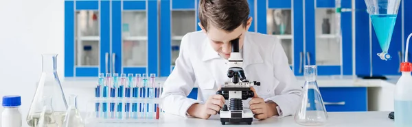 Menino de casaco branco olhando para o microscópio perto de frascos e tubos de ensaio em laboratório químico, banner — Fotografia de Stock