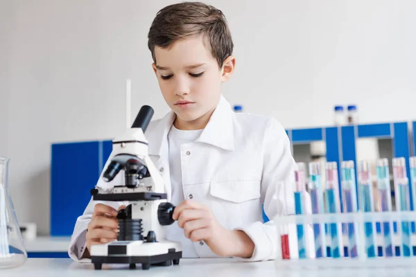Мальчик-подросток в белом халате рядом с микроскопом и размытыми пробирками в лаборатории — стоковое фото