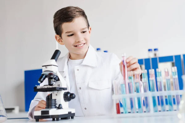 Niño preadolescente sonriente en capa blanca sosteniendo tubo de ensayo cerca del microscopio en laboratorio químico - foto de stock