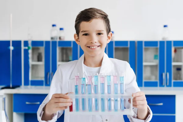 Alegre chico de capa blanca mirando la cámara cerca de tubos de ensayo en el laboratorio - foto de stock