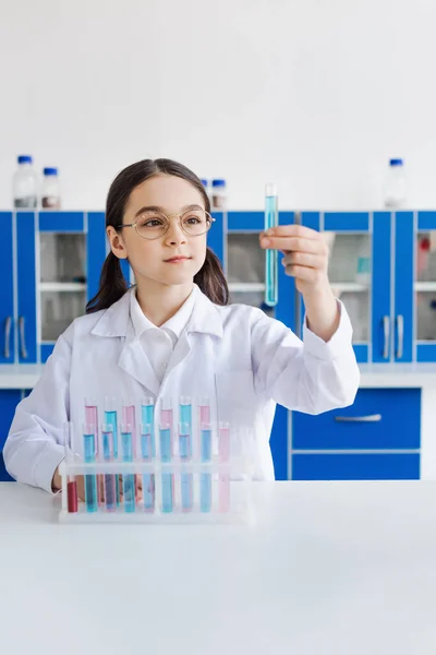 Preteen girl in eyeglasses and white coat looking at test tube in chemical laboratory — Stock Photo