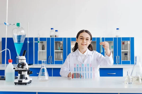 Fröhliches Mädchen mit Brille und weißem Kittel, das Reagenzglas hält, während es neben Mikroskop und Fläschchen sitzt — Stockfoto